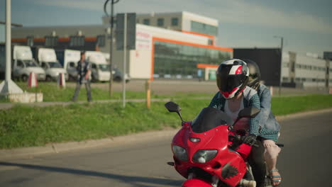 two women riding a red power bike, with the rider s shirt fluttering in the wind as her friend holds on from behind, with a blur view of cars, building, and a person walking along a sidewalk