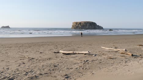 a man with long red hair spins on the beach sands with massive rocks and waves behind him