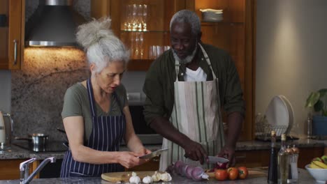 sick mixed race senior couple wearing aprons chopping vegetables together in the kitchen at home