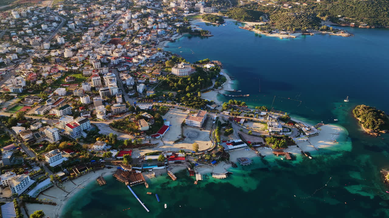 Aerial drone view of Ksamil town, it's dense cluster of hotels and houses, and the iconic bright blue bays with floating platforms and beach clubs