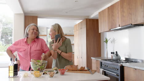 preparing salad, two multiracial senior female friends laughing and using smartphone, at home