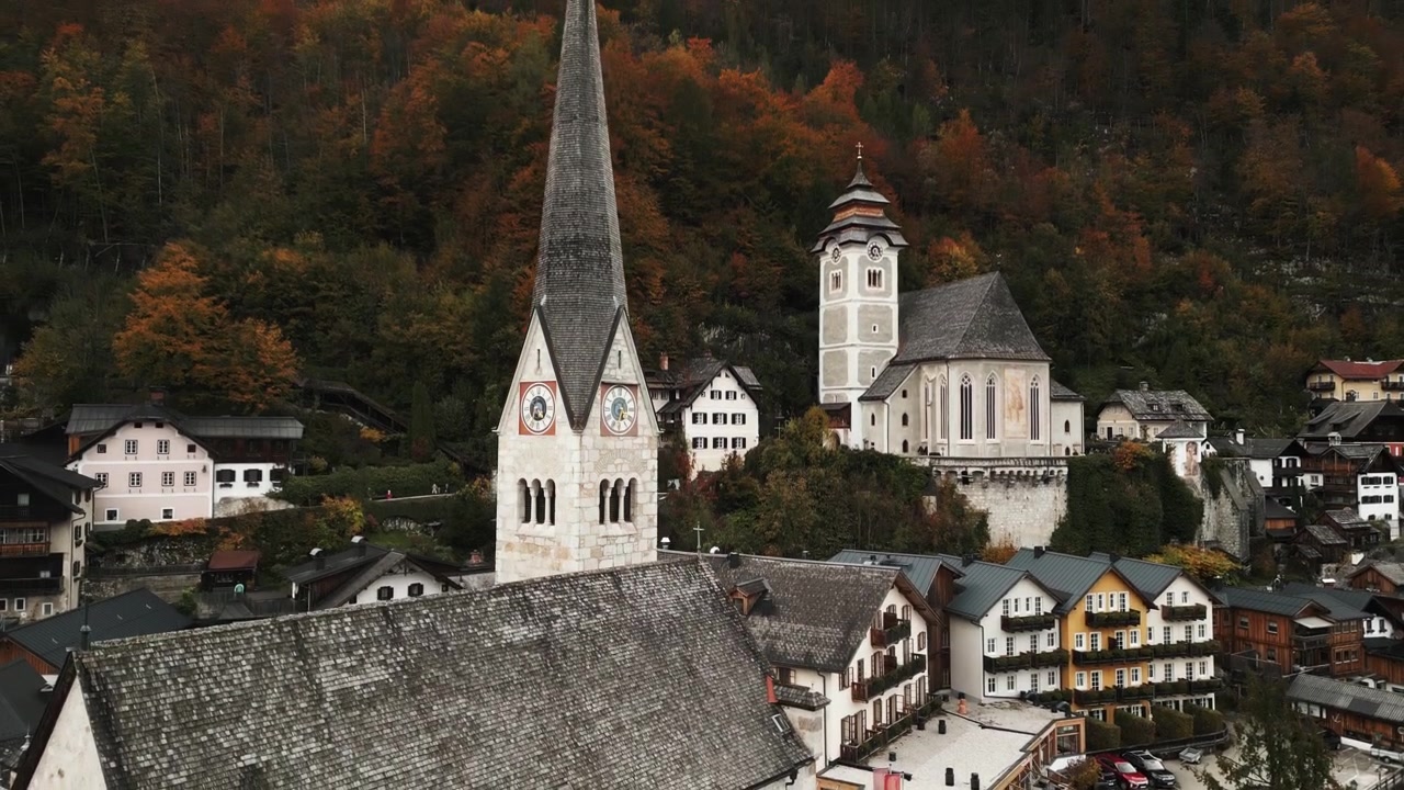 Ein malerisches Alpendorf präsentiert seine historischen Kirchen inmitten leuchtender Herbstbäume, was einen Kontrast zur traditionellen Architektur und Bergkulisse bildet