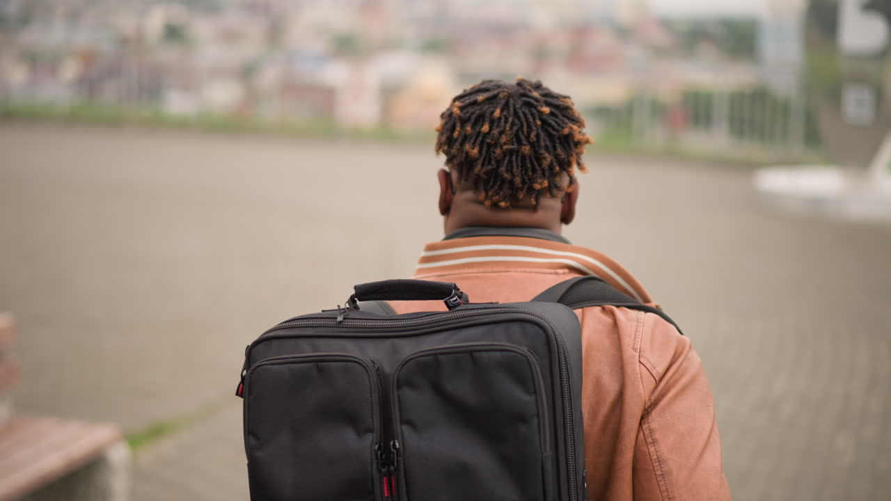 Black man with backpack in plaza walking away from camera, wearing headphones and jacket, rear view closeup with blurred city skyline and empty bench, overcast light creating moody yet hopeful