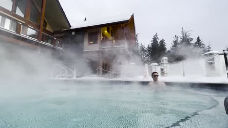 wind blowing steam off of a hot pool swirling at halcyon hot spring resort in british columbia, canada with a young white male with glasses relaxing in the steam and the resort in full view