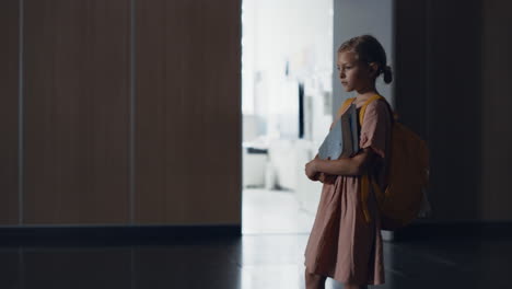 Worried-little-schoolgirl-stop-in-school-hallway-alone.-Student-holding-books.