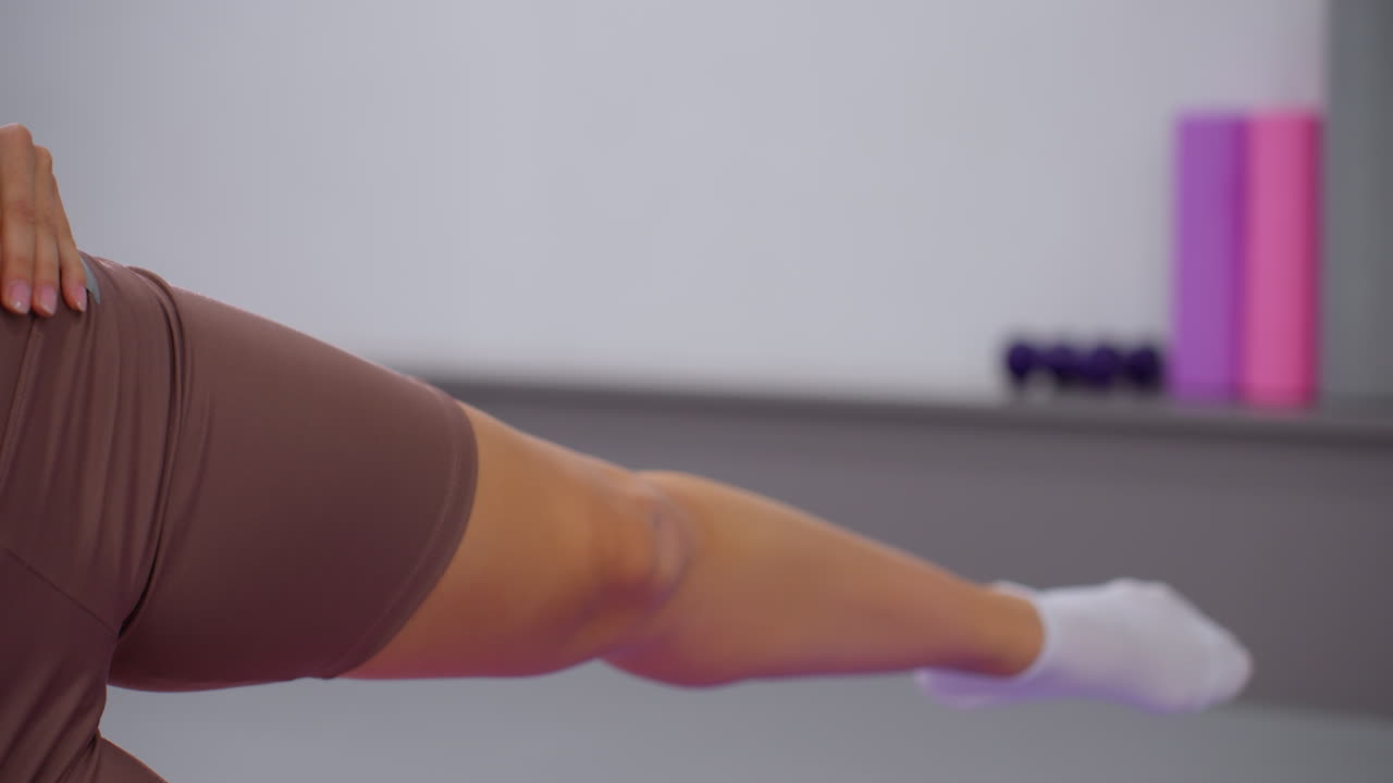 갈색 스포츠 옷을 입은 여성이 체육관에서 다리를 펴고, 하 양말을 입은 발, 약간 높이, 체육관 장비와 파스텔 색의 세부 사항으로 흐릿한 배경