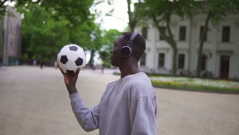 A-Stylish-Man-In-Casual-Clothes-And-Black-Headphones-Is-Spinning-A-Football-Ball-Above-Him-While-Standing-On-City-Street-Alone