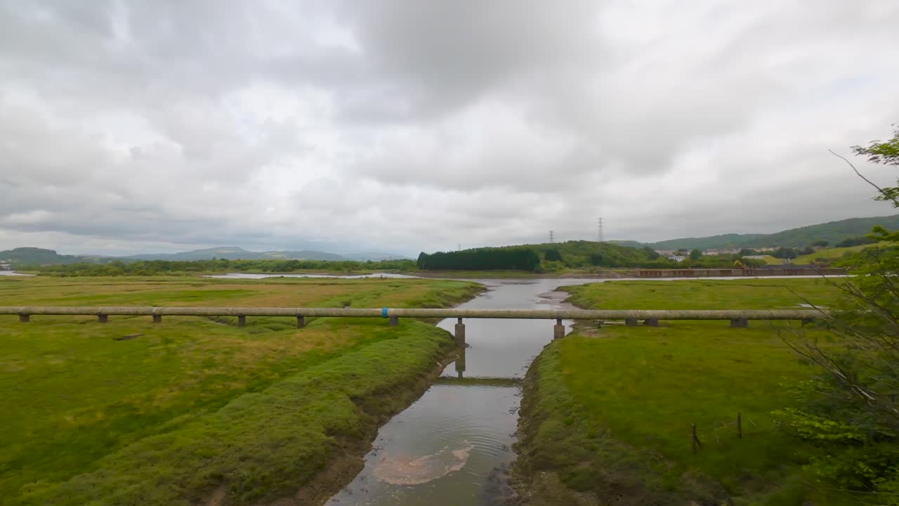 Slow Motion Wide Angle from Trees Revealing Natural Gas Pipeline Crossing Polluted River in Dense Marshland with Forest and Pylons in Background. Energy Infrastructure Footage.