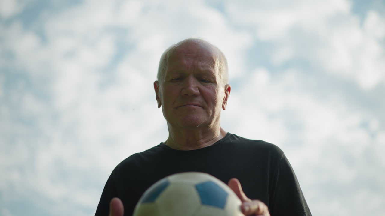 An elderly man holds a soccer ball in his hands, carefully rotating it as he lifts it up, focused and engaged, with a cloudy sky in view