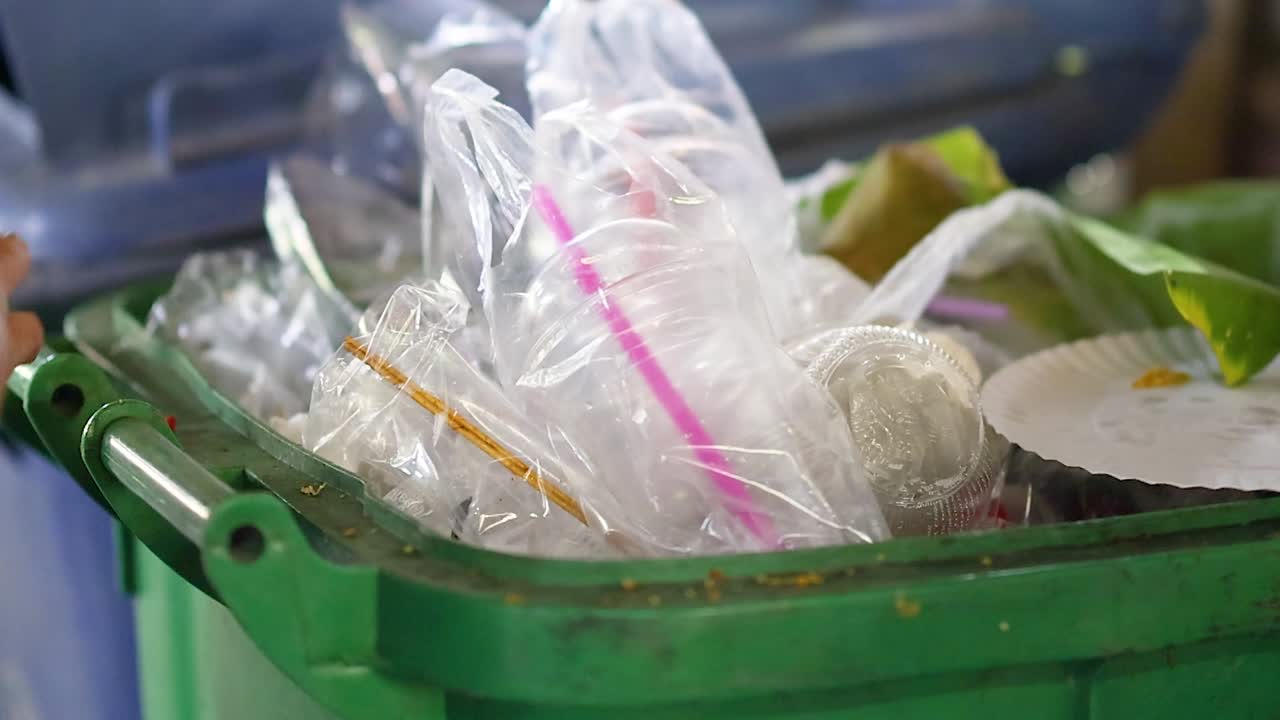 A person's hand places plastic bags and food containers into a green waste bin