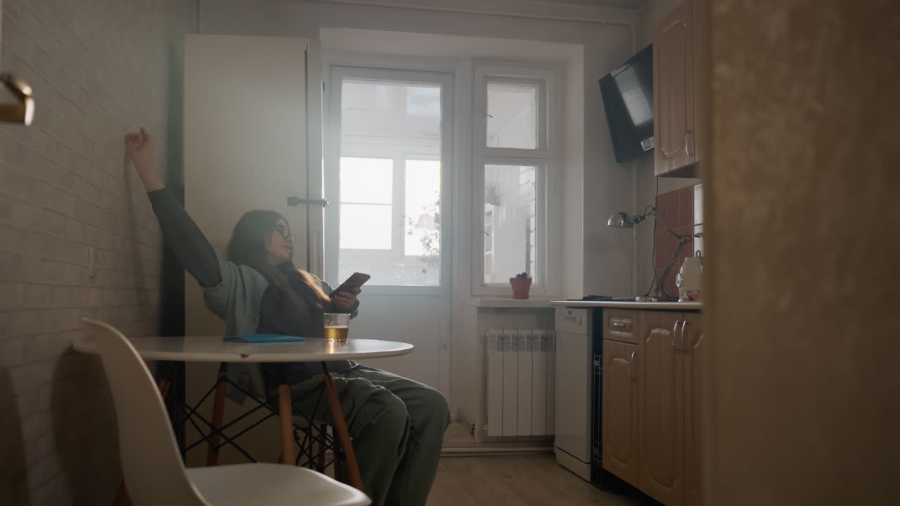Mutfak masasında telefonla oturan genç bir kadın. Pencereden süzülen sabah ışığı. Mesajlar arasında geriniyor, kupasından yudumluyor. Üzerinde rahat ev kıyafetleri ve terlikler var. Samimi küçük bir daire sahnesi, düşünceli ve rahatlamış bir atmosfer.