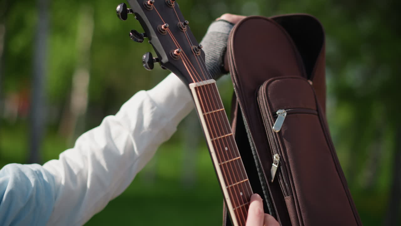 hands unzipping acoustic guitar case closeup of headstock and fingers preparing instrument, soft sunlight through green trees, denim sleeve and fingerless glove, songwriter tuning before practice