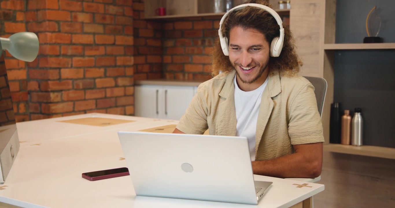 Homem na casa dos 30 anos participando de reunião online na mesa, colocando fones de ouvido, digitando em laptop prateado para trabalhar.