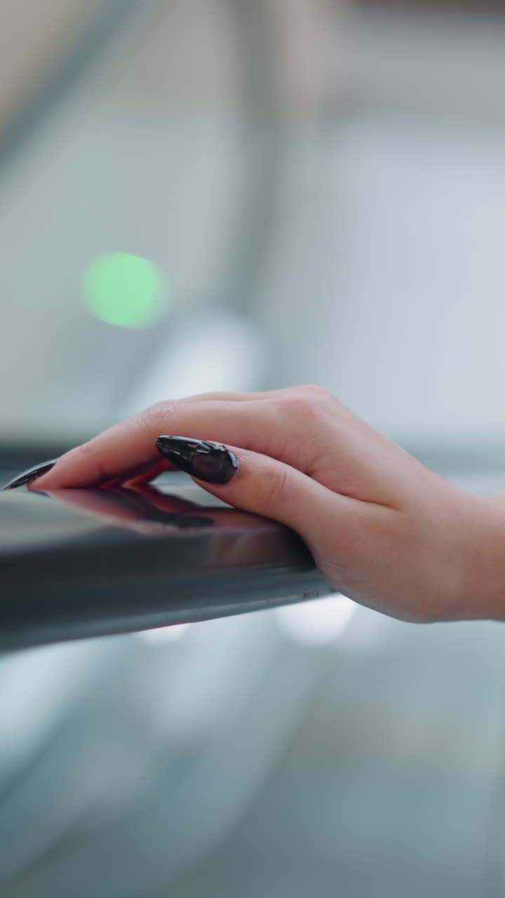 primer plano de la mano de la dama con largas uñas pulidas negras descansando en el riel de la escalera mecánica mientras asciende, el fondo presenta luces verdes bokeh suaves en un entorno interior moderno