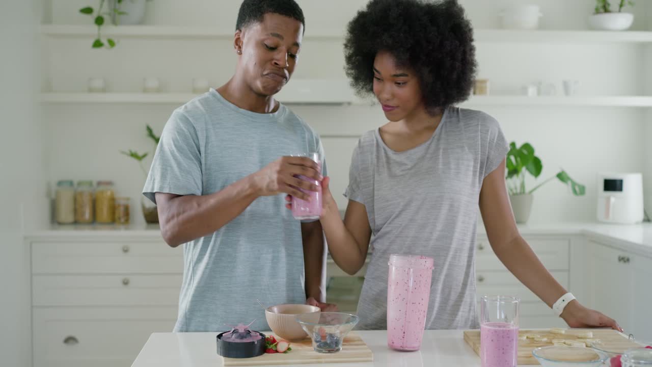 Afroamerikansk par hælder lyserød smoothie fra blenderen ved køkkenbordet, tilbyder et glas og deler.
