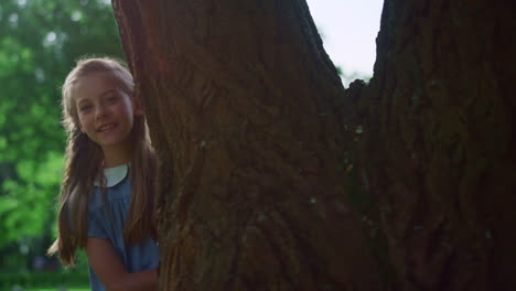 Playful girl looking out large tree trunk alone. Child peek hide on sunny day.