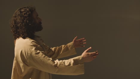 Portrait-Of-Man-Wearing-Robes-With-Long-Hair-And-Beard-Representing-Figure-Of-Jesus-Christ-Raising-Hands-In-Prayer