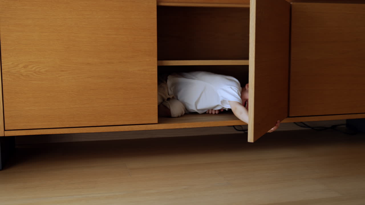 Little kid lies inside the commode hiding in it. Laughing boy opens and closes the door playing hide-and-seek