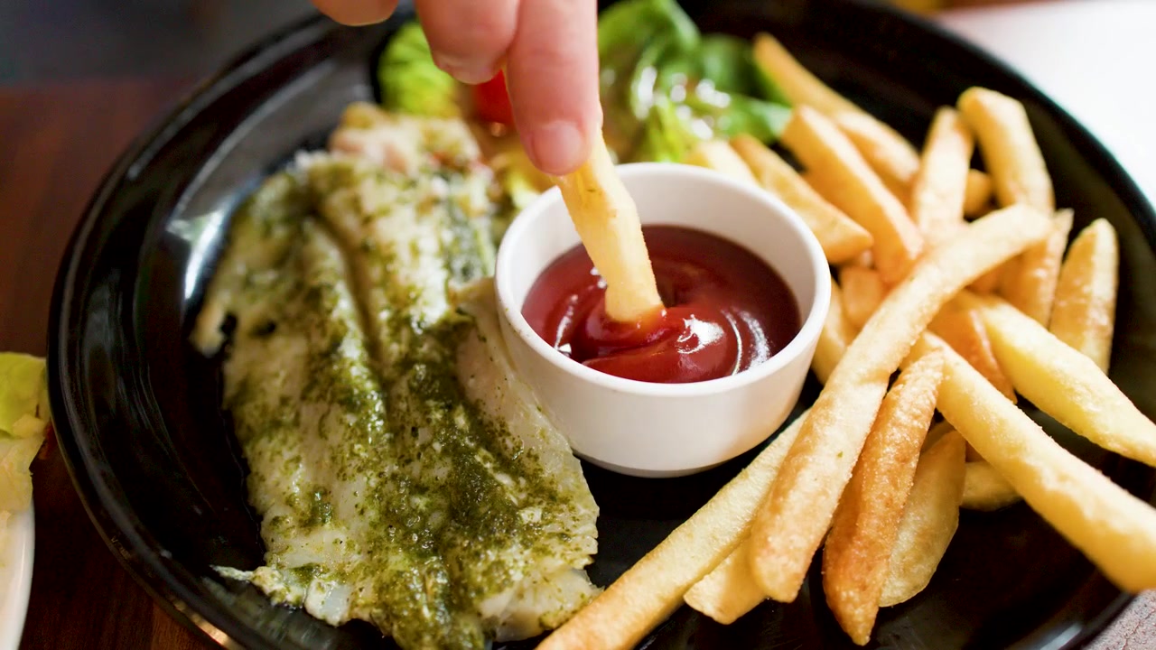 Primo piano di una mano che intinge una patatina fritta dorata nella salsa ketchup di pomodoro rossa.