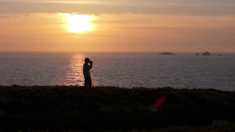 Silhouette-of-boy-with-camera-atop-coastal-cliff-with-epic-sunset-background