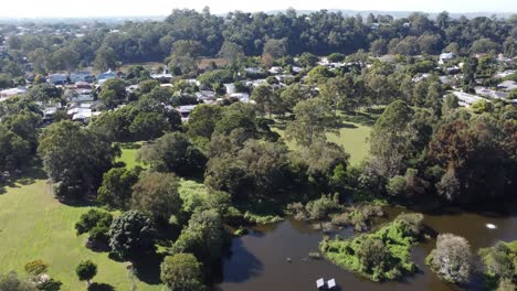 Drone-flying-over-a-small-lake-near-a-green-park-towards-residential-subdivision-in-Australia