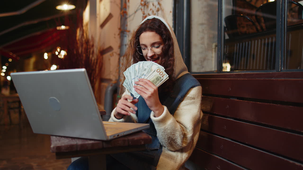 Happy rich girl using laptop winner victory holding fan of cash money in dollar banknotes in cafe