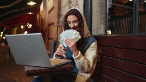 happy rich girl using laptop winner victory holding fan of cash money in dollar banknotes in cafe
