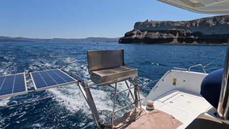 POV-looking-out-the-back-stern-of-a-yacht-with-a-BBQ-and-solar-panels,-sailing-past-the-island-of-Thera-Santorini-in-Greece-on-a-summers-day