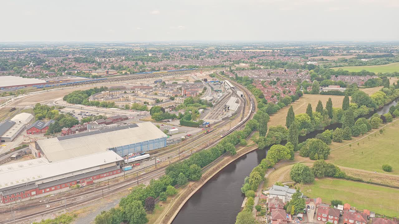Gradual aerial push-in over York, England, revealing the River Ouse curving alongside railway lines, industrial buildings, green fields and residential neighborhoods