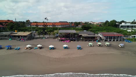 Morning-People-Relaxing-on-Seafront-Deckchairs-Lying-Under-Parasols-Near-707-Beachberm-Beachfront-Cafe-In-Batu-Belig-Beach,-Bali-Indonesia---Aerial-Rising-Revealing-Shot