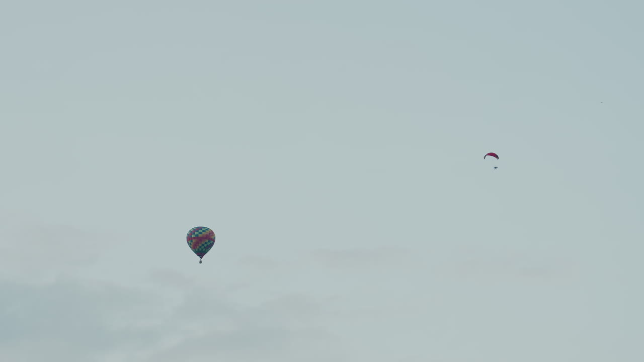 distant silhouettes of paraglider pilot and colorful hot air balloon drifting gently across pale sky evoking sense of freedom calm silent aerial journey above soft cloud patterns
