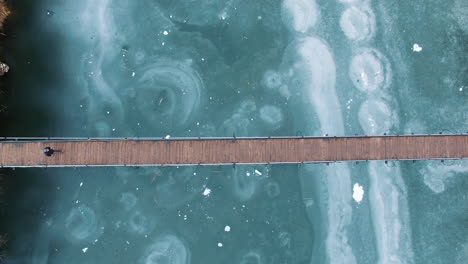 AERIAL:-A-man-on-crutches-crosses-a-wooden-bridge-over-a-frozen-lake