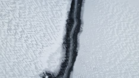 Drone-top-down-follows-exposed-waterway-edge-splitting-icy-lake-and-windswept-jagged-pattern