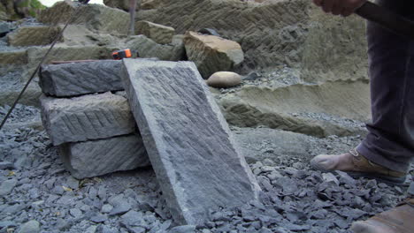 handheld wide shot of a craftsman chipping a stone slab, with a hand pick, shot outside in the city of ancud, chiloe island