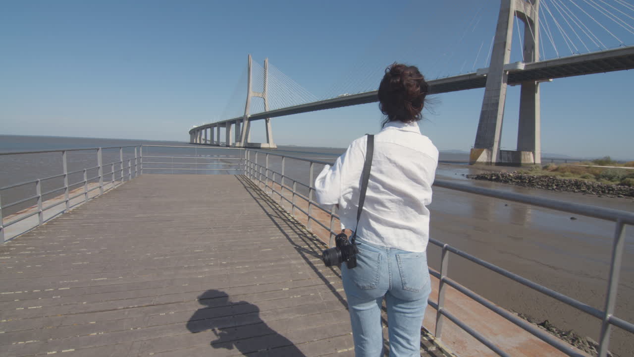 Follow shot of a young woman walking down the pier and enjoying the amazing view of the river with a bridge. Low tide.Tagus river. Contemplation in solitude. Architectural wonder