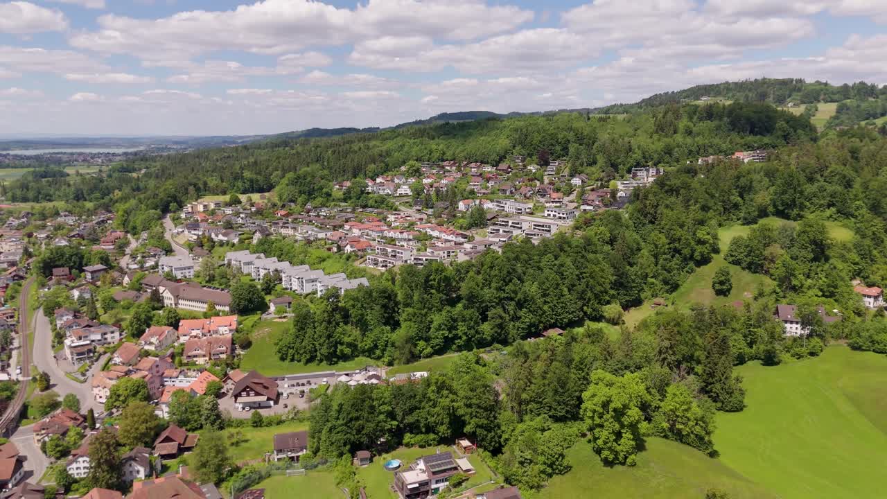 Aerial establishing shot of luxury swiss city on hillside with green trees. CLouds at sky in summer. Wide shot. Homes and houses in noble district.