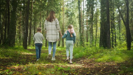 back view of woman walking in forest with children, holding girl hand as she holds grass, while boy walks beside them, surrounded by lush green trees, soft light, and peaceful woodland scenery