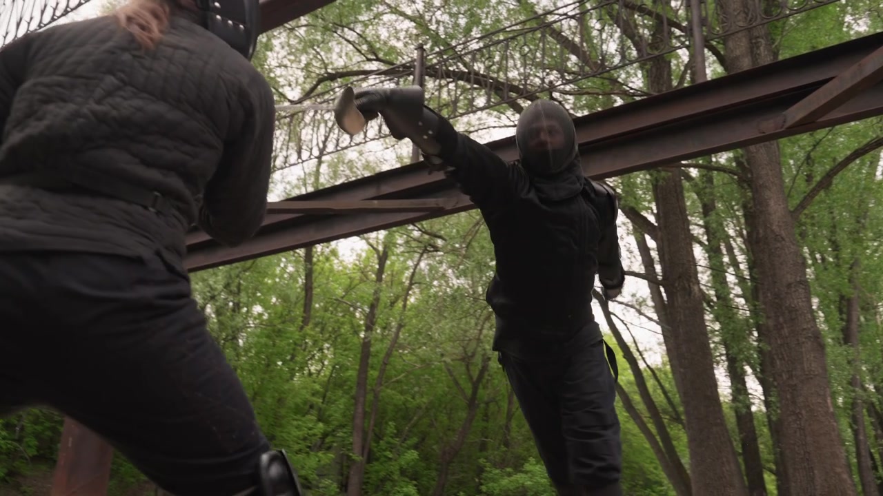 Two people in black protective gear engage in sword fight under metal beams with trees visible. One person wields sword while other prepares to defend both wearing padded clothing