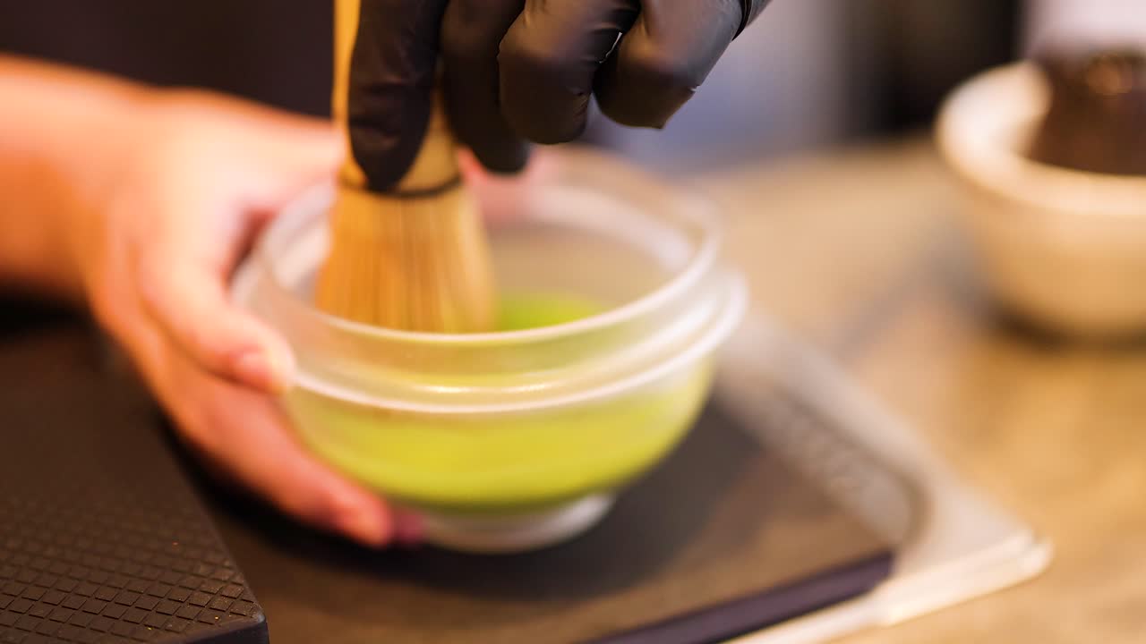 Hands whisk matcha in a bowl with precision. Soft lighting enhances the serene atmosphere of this traditional tea-making process