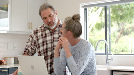 mature couple using laptop in kitchen, discussing plans with thoughtful expressions
