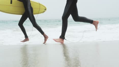 Unterer-Abschnitt-Von-Zwei-Männlichen-Surfern,-Die-Mit-Surfbrett-Am-Strand-Laufen-4k