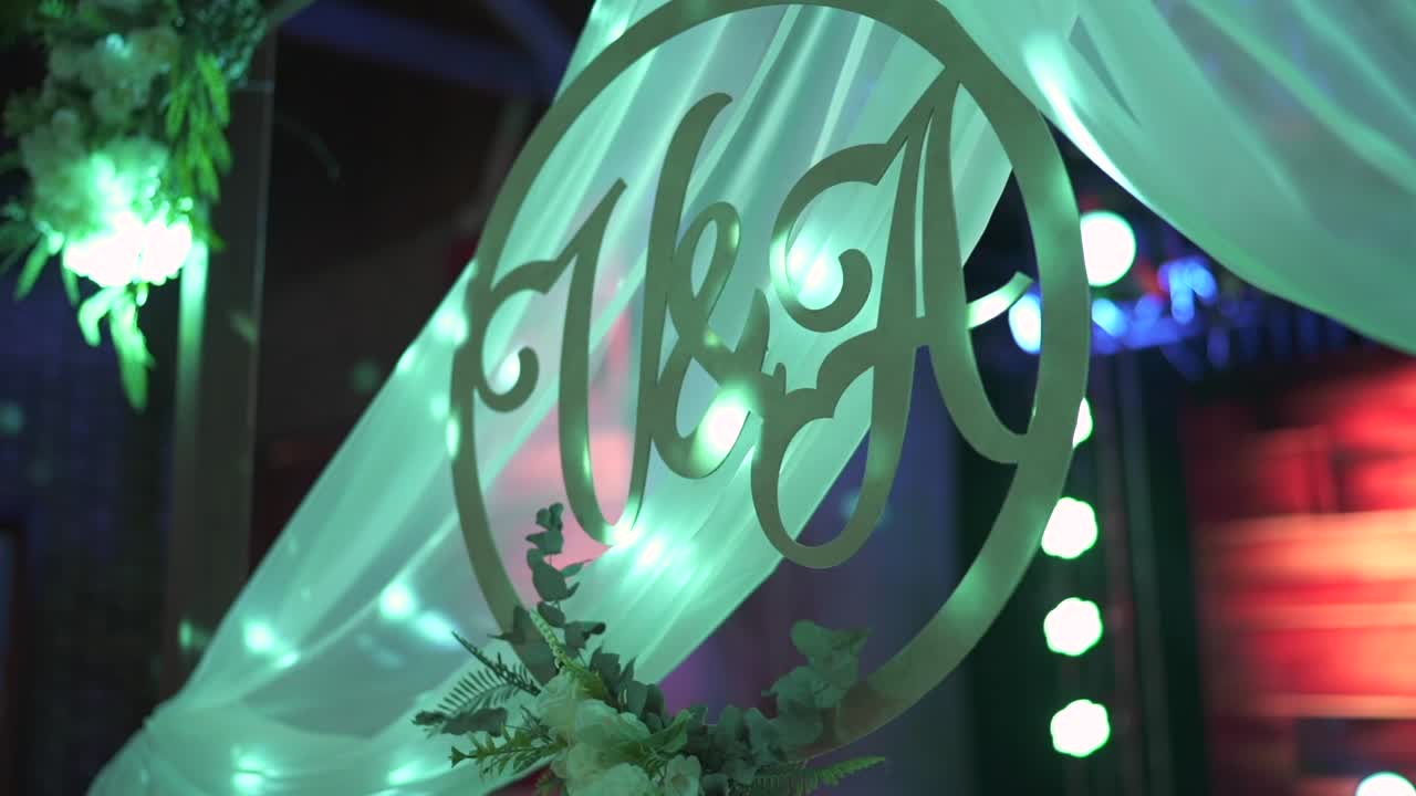 Artistic close-up of a wooden circular sign displaying the initials V and A, symbolizing the bride and groom, surrounded by flowers and white drapery under romantic green lighting at wedding reception
