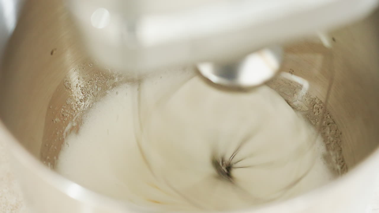 Close up of electric stand mixer spinning flour and liquid ingredients in shiny stainless steel bowl, creating dough or batter in motion, ideal for baking and kitchen preparation concepts
