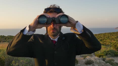 hombre en traje elegante con maleta mirando a través de binoculares en la parte superior de un acantilado en una isla