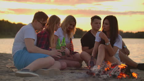 five young people are sitting in shorts and t-shirts around bonfire on the sand beach. they are talking to each other and drinking beer at sunset.