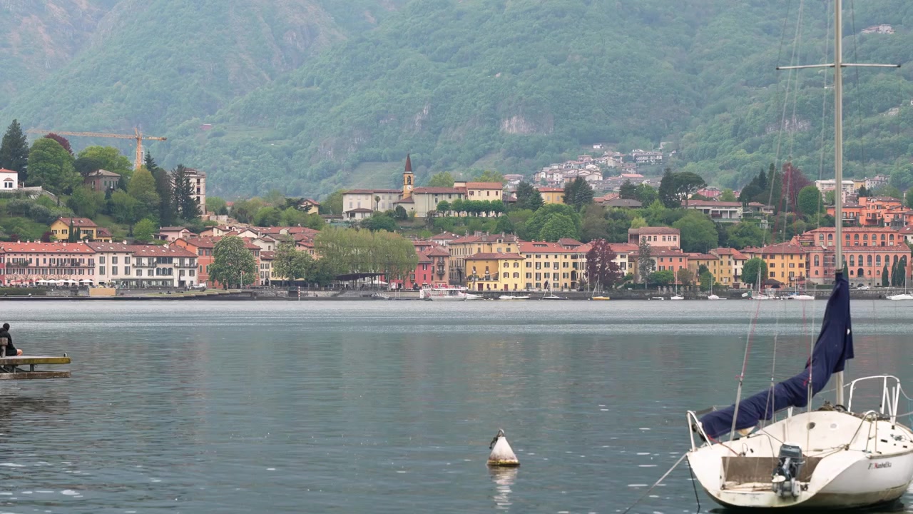 Ampia inquadratura serena della città di Lecco sulla sponda del Lago di Como, Italia, con edifici colorati, colline lussureggianti e una barca a vela ormeggiata sotto una luce diurna soffusa, che riflette un paesaggio lacustre tranquillo.