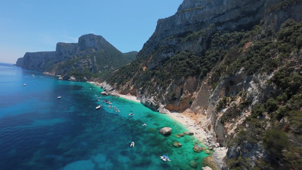 Turkist vand møder tårnhøje kalkstensklipper, mens en FPV-drone flyver tæt langs den barske kyst mod Cala Goloritze, Baunei, Sardinien, Italien