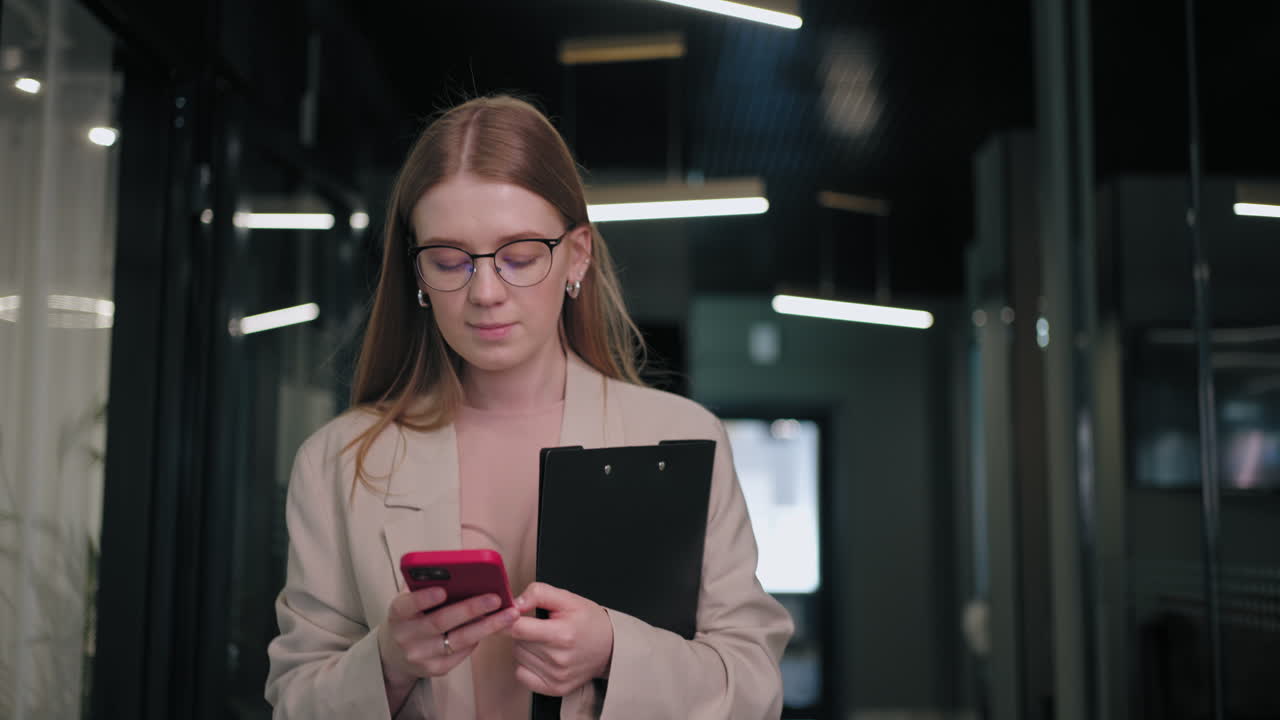 mujer de negocios sonriente mirando el teléfono celular en el interior. mujer de negocios sorprendida leyendo un mensaje en el teléfono móvil en el pasillo de la oficina. mujer mirando la pantalla del teléfono inteligente en el centro de negocios. oficina caminando