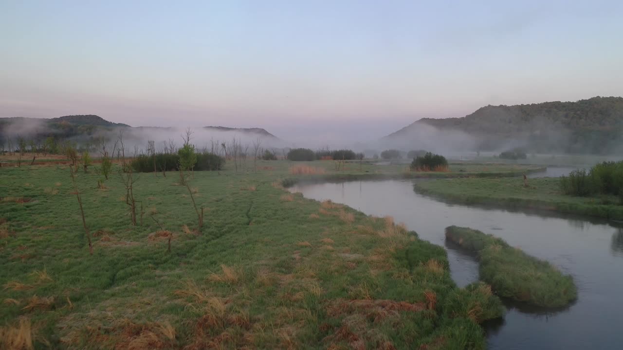 Serene aerial view of a mist-covered river flowing through the quiet countryside at dawn. Soft morning light filters through, creating a peaceful and tranquil atmosphere