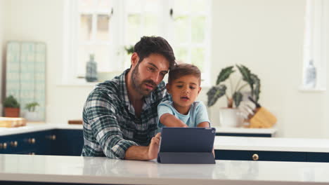 Padre-En-Casa-En-La-Cocina-Con-Su-Hijo-Transmitiendo-O-Jugando-En-Una-Tableta-Digital